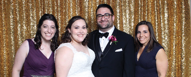 A bride and groom with two women on either side smile with gold sparkly curtains in the background